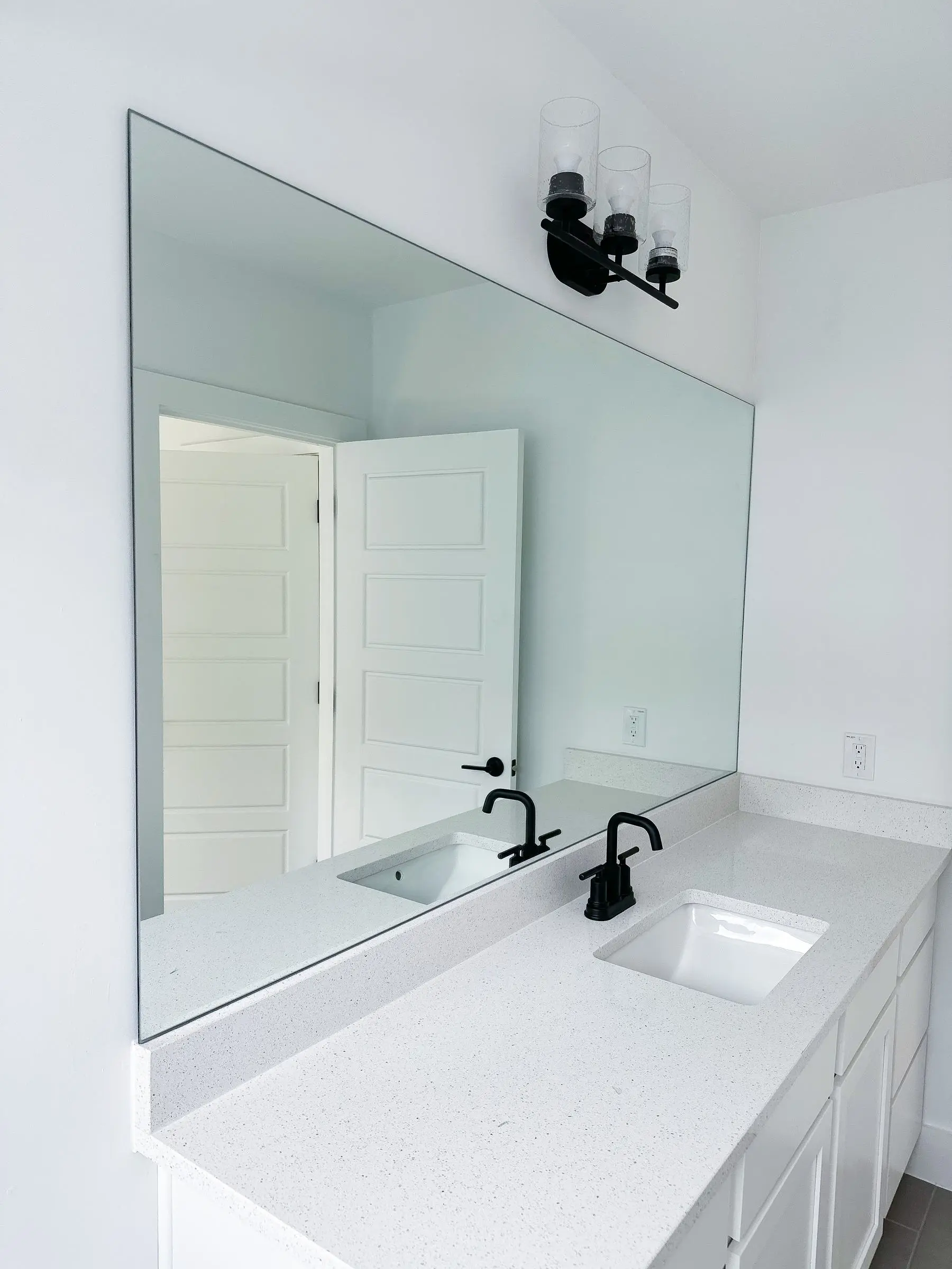 A large frameless mirror cut to the wall above a white quartz double vanity, with black faucets and a black three-globe light.