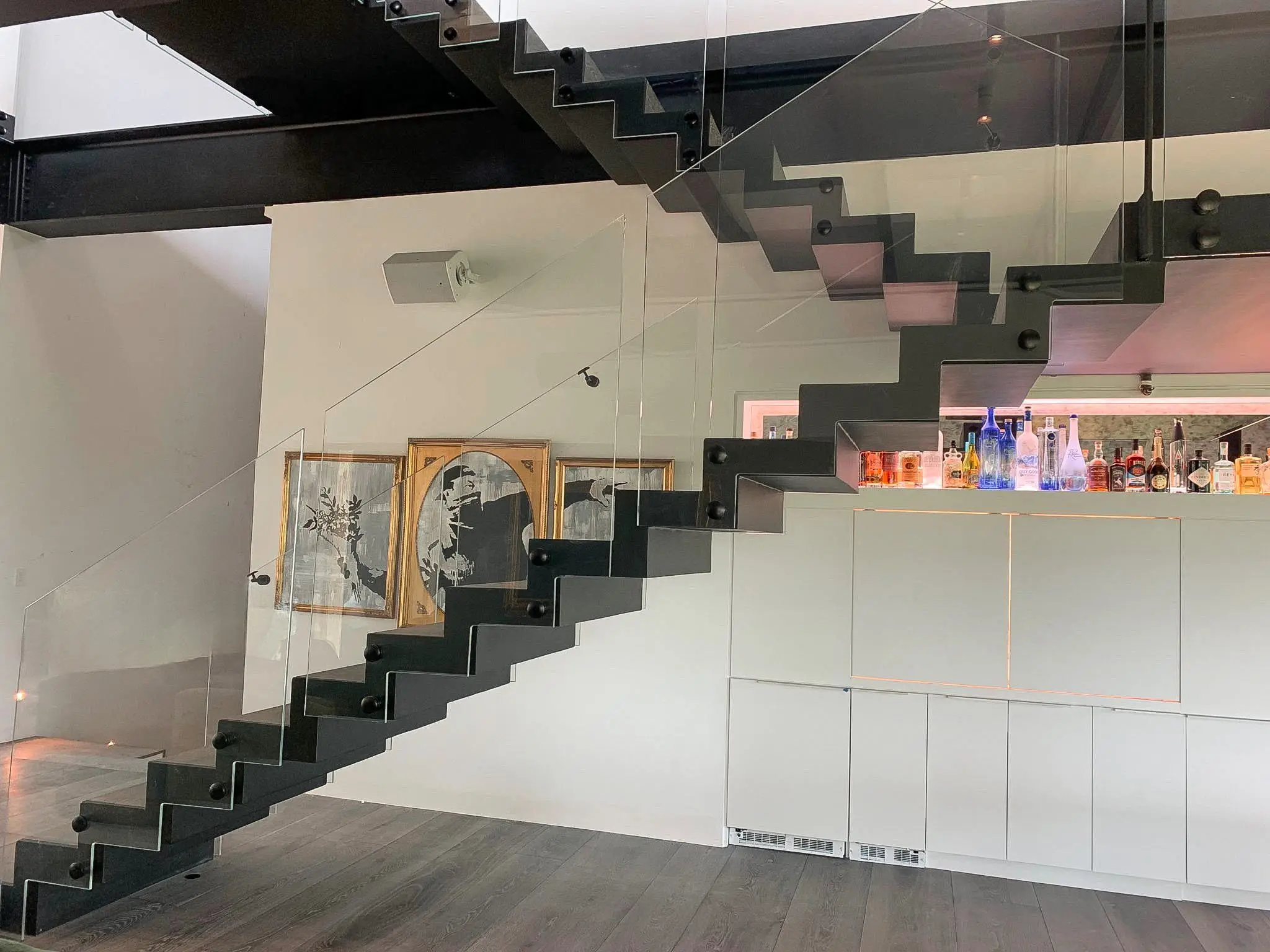 A frameless glass railing on a black floating steel staircase, with a framed picture wall behind and a backlit bar beneath the stair.