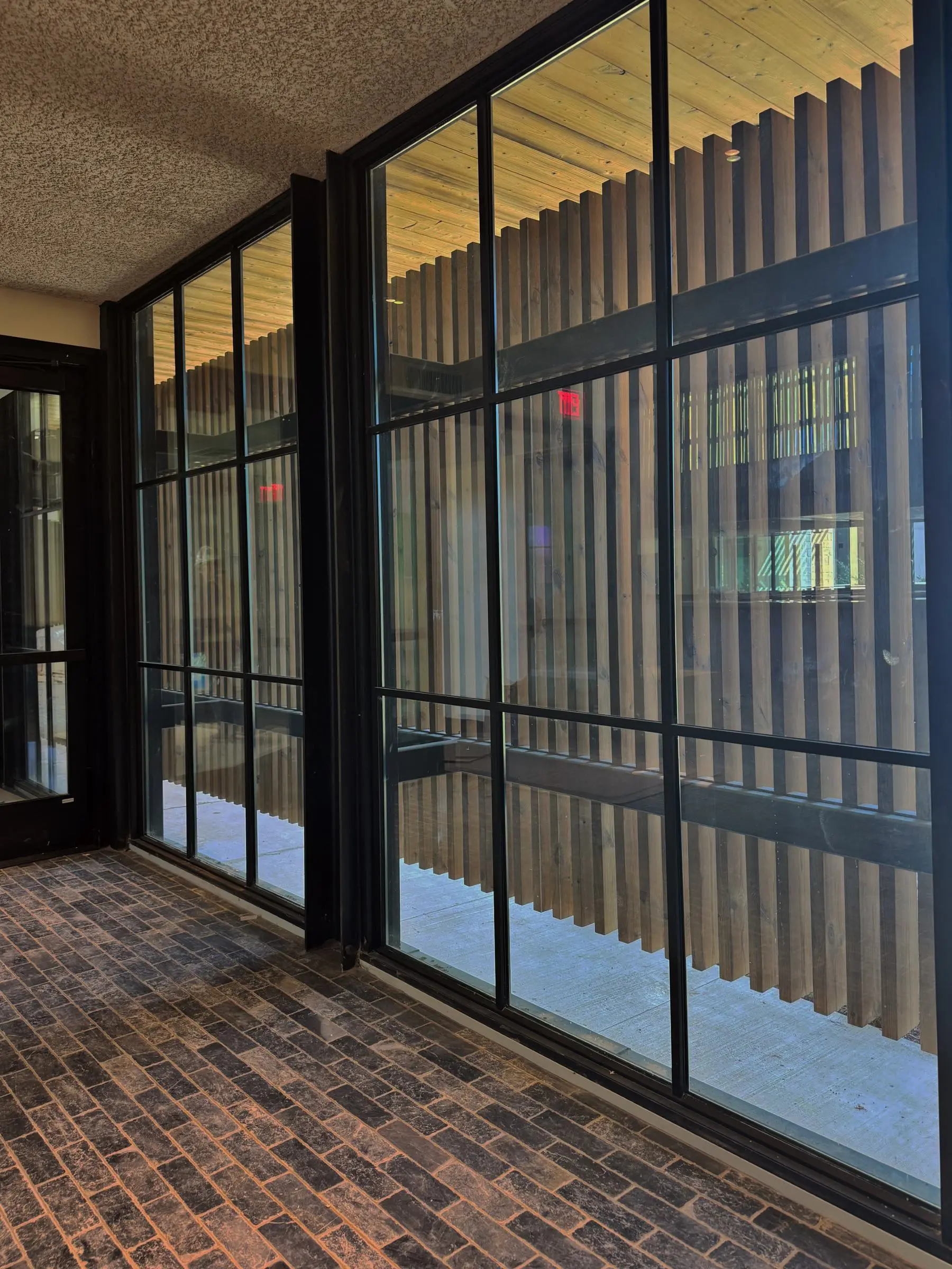 A black-framed interior window wall along a brick-floored corridor, looking out onto a slatted wood screen.