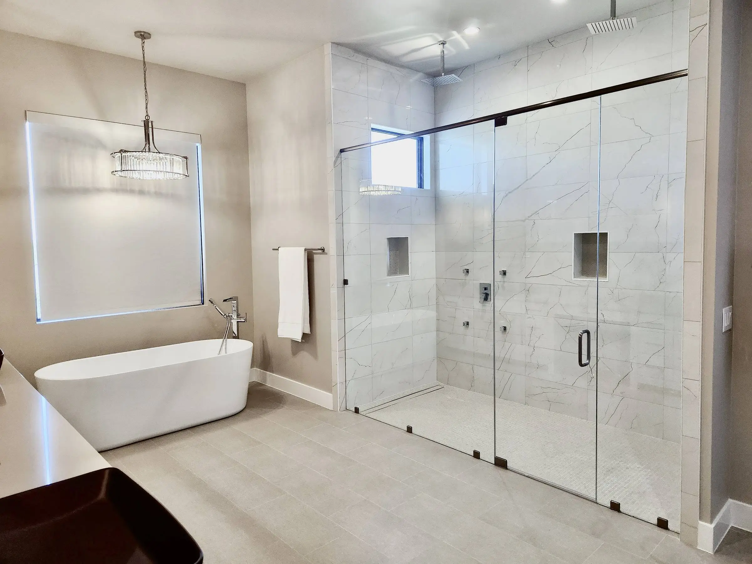 A bathroom with a large frameless glass shower enclosure along one wall, a freestanding white tub under a window, and a cut-glass pendant light.