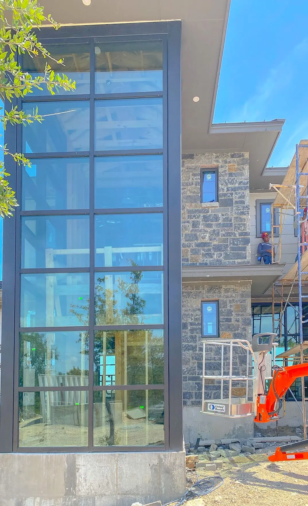 A tall multi-story window wall being installed on a stone building, with scaffolding, a boom lift, and an installer working on the upper level.
