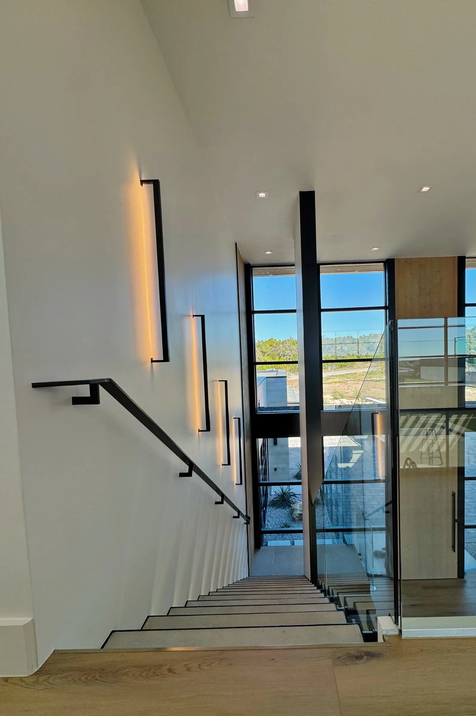 A staircase beside a two-story black-framed window wall looking out over Hill Country scrub, with a frameless glass railing at the landing.